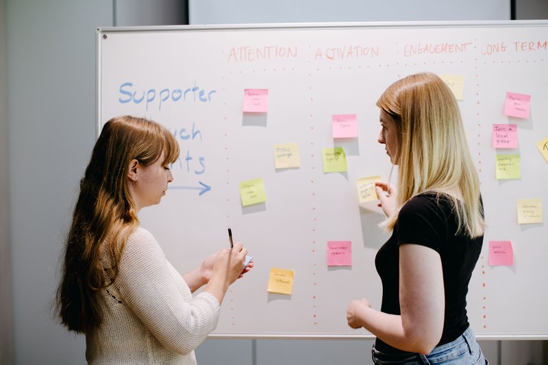 Two women use sticky notes to add ideas to a whiteboard